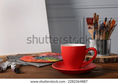 Easel with blank canvas, cup of drink and different art supplies on wooden table near grey wall