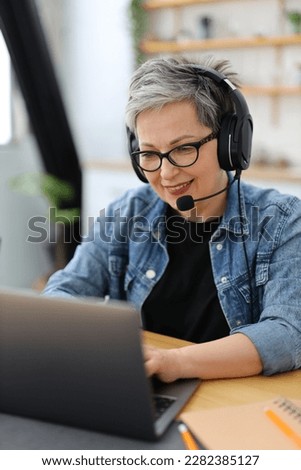 Customer service, smiling mature woman in glasses and headphones in front of a laptop. Royalty-Free Stock Photo #2282385127