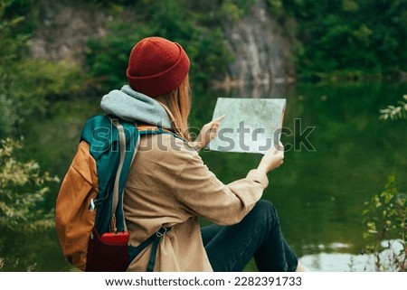 Young lady traveler in a hat sits on a near a lake with a paper map and looks into the distance. Mountains and lake on the background. High quality photo
