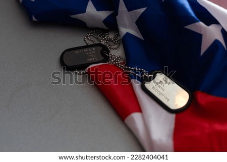 Closeup of a dog tag with the text thank you veterans engraved in it, next to a flag of the United States background.