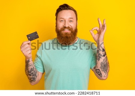 Photo of young worker hipster man wear green t-shirt okey symbol cool tattoos hold debit card wireless payment ad isolated on yellow color background