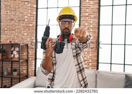Young hispanic man with beard working at home renovation pointing with finger to the camera and to you, confident gesture looking serious 
