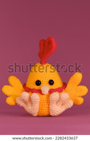 Amigurumi chicken doll on pink background. Soft DIY toy made of cotton and natural wool. Orange cockerel with bright red comb on head and beak crocheted, handmade art. Front view.