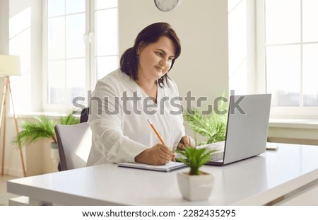 Overweight brunette young woman using laptop in modern office. Focused plus size woman reviewing financial documents and making notes on paper. Financial manager, bookkeeper, accountant Royalty-Free Stock Photo #2282435295