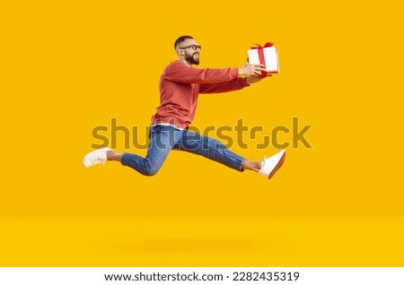 Charming smiling young man in glasses wearing red sweatshirt and jeans jumping high holding gift smiling isolated on yellow background. Holiday gifts, discounts, sales, marketing, good offer concept.