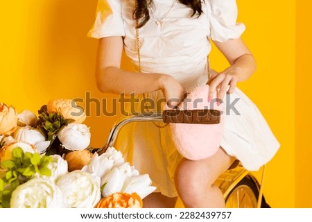 Cute stylish brunette girl in a fancy short white dress and bright makeup is sitting on a bike with flowers and searching or looking for something in her handbag. Close-up. Isolated on yellow.