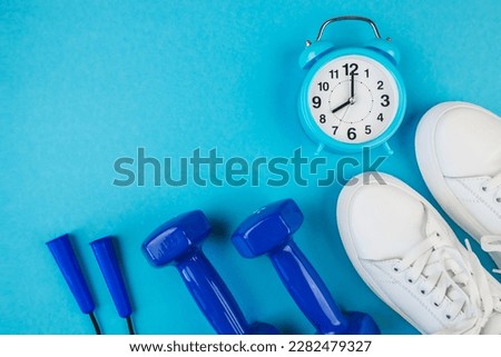 The concept of fitness, sports, weight loss. Top view photo of white sports shoes, blue alarm clock, jump rope and blue dumbbells on isolated pastel blue background with empty space