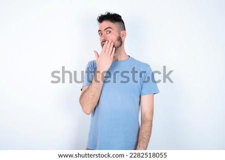 Young caucasian man wearing blue t-shirt over white background covers mouth and looks with wonder at camera, cannot believe unexpected rumors.