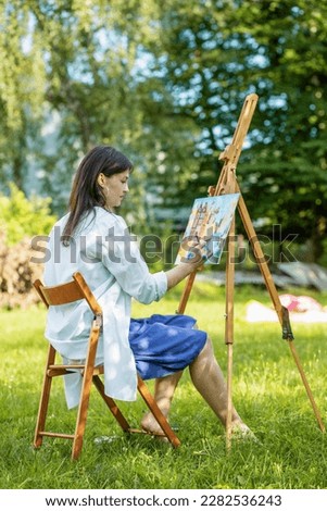 Young female artist paints picture in summer park in sunlight. Art and hobby concept. Back view