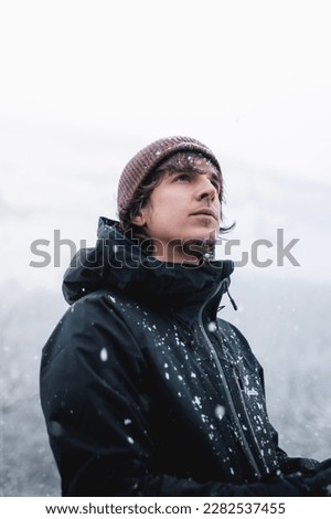 A young man with wool red beanie and black jacket under the snow flakes in winter.