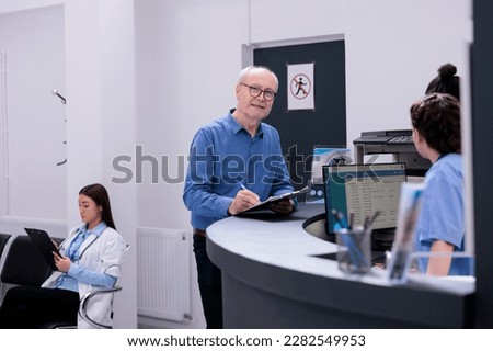 Senior patient standing at hospital reception signing medical papers before start examination with physician medic. Assistant prescribing medication to help manage the man chronic pain in lobby