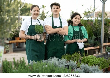Multiracial group of successful confident young floriculturists engaged in growing ornamental potted plants standing in greenhouse. Smiling young woman holding pot of fragrant rosemary Royalty-Free Stock Photo #2282553331