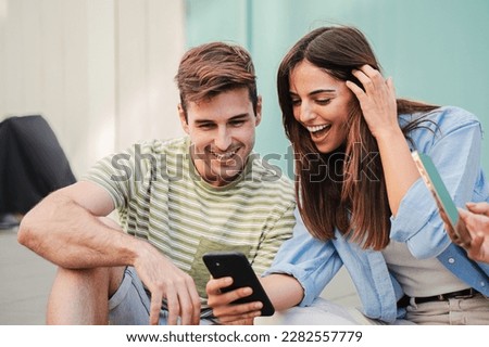 Two teenage happy friends watching funny videos browsing on internet app with a smartphone and smiling together. Caucasian couple using a cell phone to post in a social media and chatting online. High Royalty-Free Stock Photo #2282557779