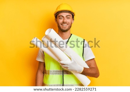 Portrait photo of young bearded man wearing yellow hard hat and holding rolls of paper, standing isolated over yellow backdrop and smiling, professional people concept, copy space, high quality photo Royalty-Free Stock Photo #2282559271