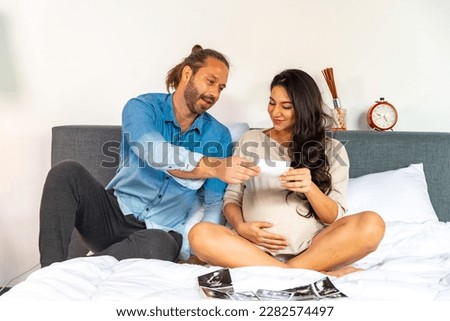 Beautiful Caucasian pregnant woman and husband looking ultrasound photo of their newborn baby together on the bed in bedroom. Family husband and wife relationship and maternity health care concept.