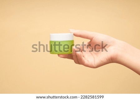 Woman's hand holds jar of moisturizing cream made of amber glass on beige background. Bottle without label with anti-aging mask, cosmetic. Concept of skincare, daily routine. Front view Royalty-Free Stock Photo #2282581599