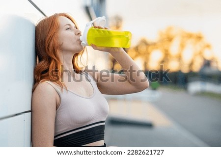 young redhead woman drinks water with her back against the wall. thirst quencher after workout