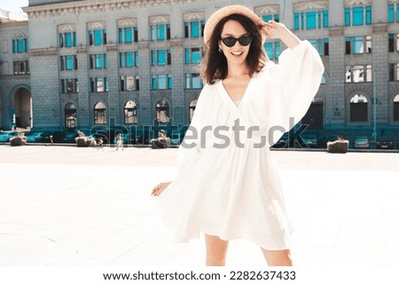 Beautiful smiling brunette model. Trendy female posing in the street background. Funny and positive woman having fun outdoors at sunset. In hat at sunny day. In white dress, sunglasses