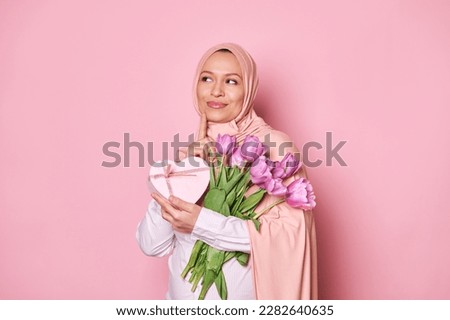 Pensive delighted Muslim pregnant woman dreamily looking aside, holding bouquet of purple tulips and heart shaped gift box, isolated on pink background. Mother's and International Women's Day concept