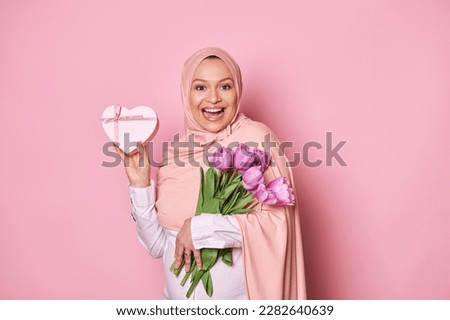 Delightful Arab Muslim pregnant woman in pink hijab, holding a bouquet of purple tulips and showing at camera a happy present in a heart shaped gift box for Mother's Day, isolated on color background