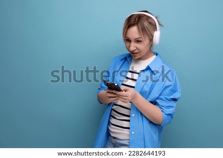 close-up photo of a bright smiling blond young woman dressed in a striped sweater and casual shirt with white wireless headphones communicates in the application in a smartphone on a blue background