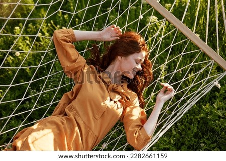 a woman is resting lying in a mesh hammock with her hands behind her head, smiling happily, enjoying a warm day in the rays of the setting sun, lying in an orange dress