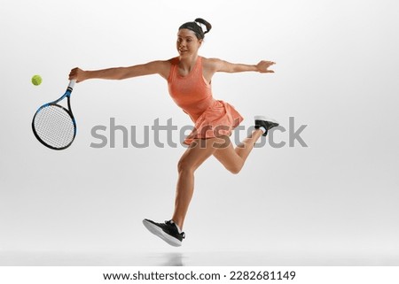 Portrait of smiling young woman in sports uniform, training, playing tennis against white studio background. Hitting ball with racket. Concept of professional sport, movement, health, action. Ad