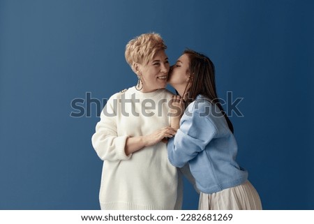 Daughter kissing mother. Happiness. Portrait of middle-aged woman and young girl posing over blue studio background. Concept of motherhood, family, mother's day, love, emotions, relationship