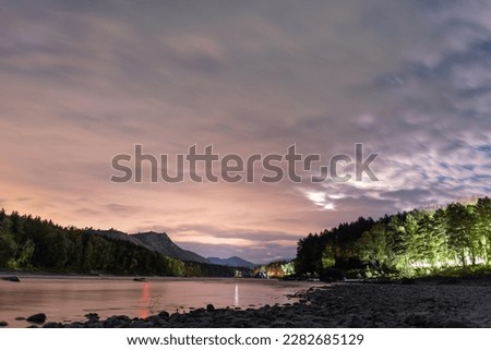 Bright night with clouds in the sky near the mountain river Katun with a red radio beacon on the rock and the light of rest houses in the forest in the Altai