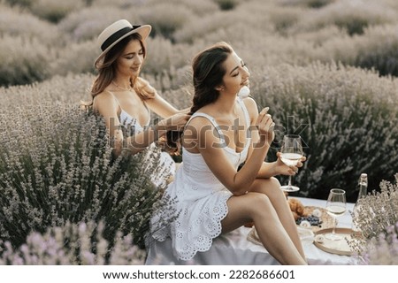 Two women enjoying a picnic with fresh fruit and wine while sitting and chatting on field of lavender. One girl braids another girl's hair. Royalty-Free Stock Photo #2282686601