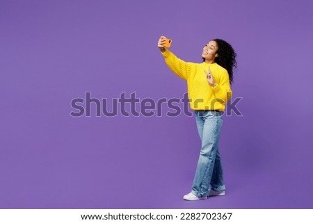 Full body young woman of African American ethnicity wear casual yellow sweater doing selfie shot on mobile cell phone post photo on social network isolated on plain purple background studio portrait