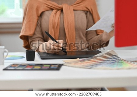 Young creative woman using graphic tablet and working with color swatch samples at workstation