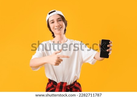 Cool teenage boy pointing at mobile phone on yellow background