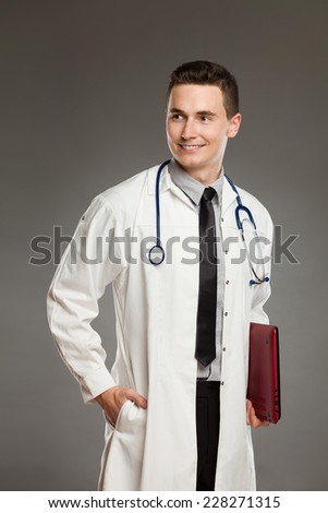 Portrait of a smiling doctor holding laptop under his arm and looking away. Three quarter length studio shot on gray background.