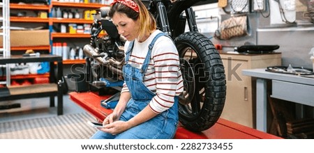 Portrait of smiling mechanic woman looking phone in her hands sitting over platform with custom motorcycle on factory