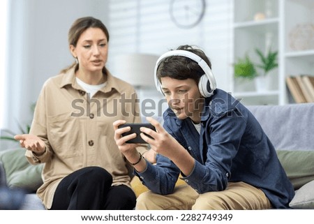Family conflict quarrel, woman mother quarrels with son, teenage boy in headphones ignores woman and plays video games on phone, family at home in living room sitting on sofa. Royalty-Free Stock Photo #2282749349