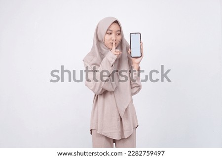 Mysterious Asian woman with brown hijab makes silence gesture presses index finger to lips shares secret with you holds mobile phone and warm jumper isolated over white wall