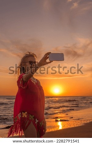 Senior woman with sunglasses taking a selfie with a mobile phone during sunset at the beach. Summer concept.