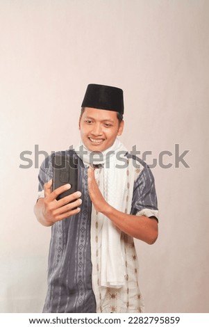 asian muslim youth in video call with cell phone saying one hand greeting, happy eid al fitr. Eid al-Fitr advertisement photo concept for Muslim holiday