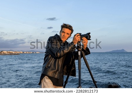 Handsome professional photographer in the spring takes pictures of the sea landscape. Conceptual photo funny meme with face to study Out door travel photography with tripod and reflex.  School photo. 