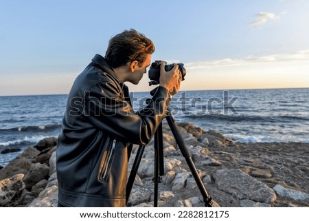 Handsome professional photographer in the spring takes pictures of the sea landscape. Conceptual photo to study travel photography out of town with tripod and reflex camera