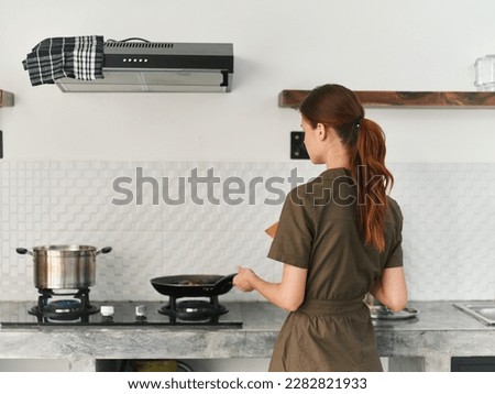 A woman at home smile in the kitchen prepares food stands with her back to the camera at the stove and fries meat and vegetables in a pan, a large kitchen in a modern style, lifestyle Royalty-Free Stock Photo #2282821933