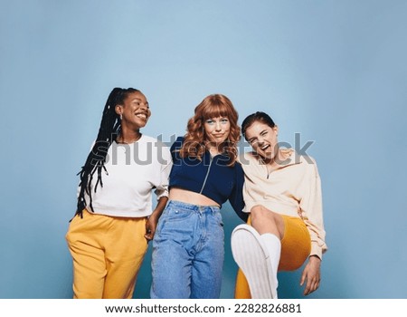Group of happy female friends having fun while standing together in a studio. Three cheerful female friends smiling and having a good time against a blue background.