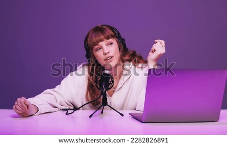 Young woman speaking into a microphone while hosting a live radio show. Female podcaster recording an audio broadcast in neon purple light. Woman creating content for her internet podcast in a studio.