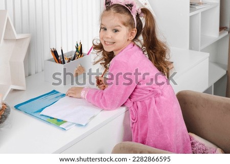 A charming overjoyed little girl in a pink pajama and robe is standing next to her desk and drawing with colorful pencils. Royalty-Free Stock Photo #2282866595