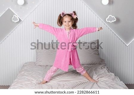 A charming little girl in a pink pajama and robe is jumping on the bed, happily throwing her hands up and laughing. Royalty-Free Stock Photo #2282866611