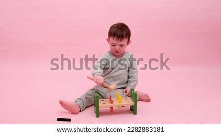 Happy toddler baby plays with a hammer and wooden nails on a studio pink background. Child boy with educational toy. Kid age one year eight months, full height