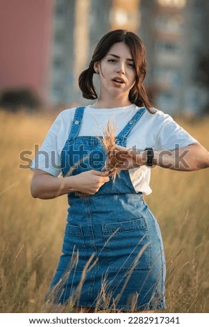 Portrait of a young beautiful dark-haired girl in a denim sundress on a summer field. Royalty-Free Stock Photo #2282917341