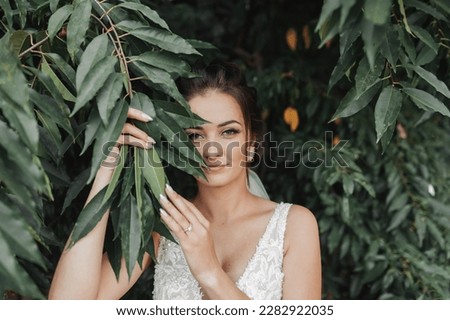Wedding photo on a green background. The bride in a white dress stands near green bushes, holds a leaf near her face and looks into the lens. Beautiful makeup. Portrait of the bride