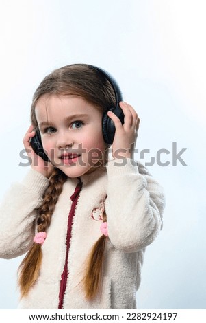Portrait of a girl in a white warm sweater on a white background with headphones on her head, the child is braided in two long braids, a cute smile of a little girl.